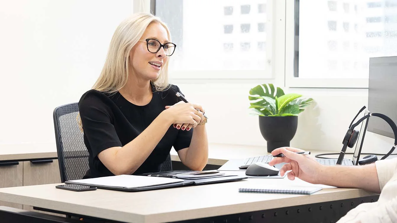 A female employee wearing a black top and glasses having a conversation with another employee at her desk.