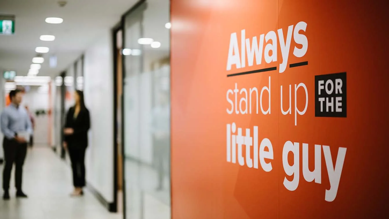 An orange wall with 'Always stand up for the little guy' displayed on it, with a male and female worker blurred in the background.