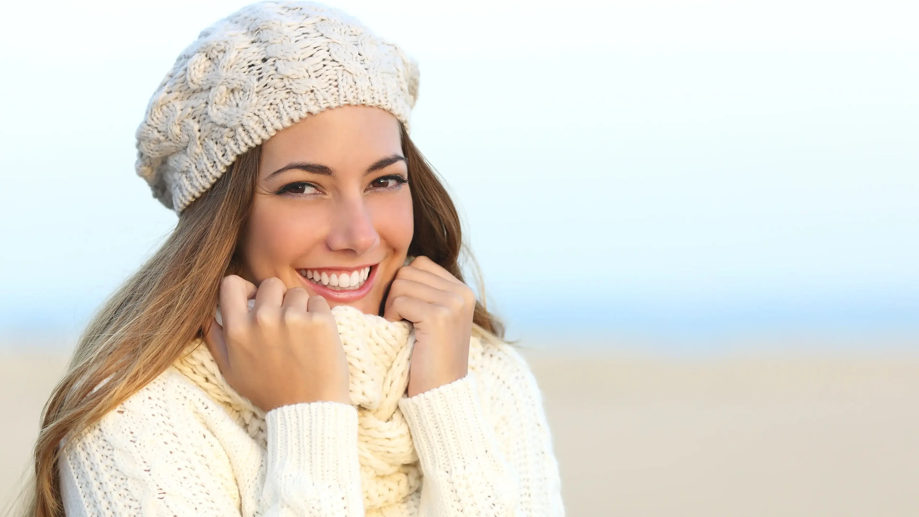 Eine lächelnde Frau mit Mütze am Strand, die ihren Hals mit ihrem Schal verdeckt.