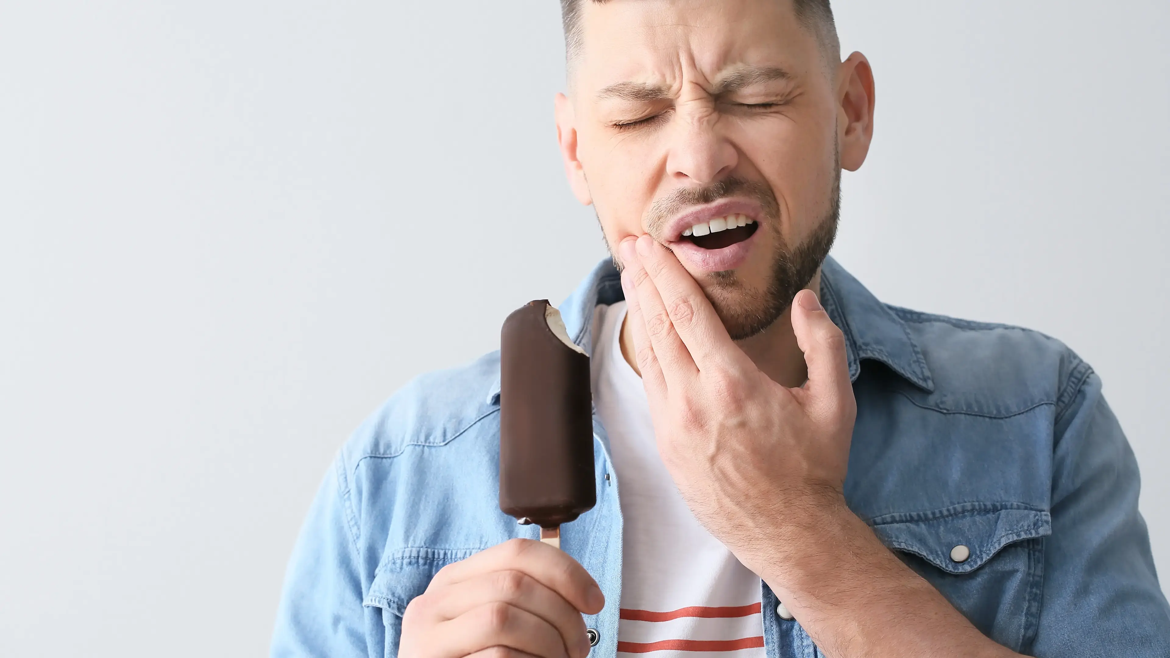 Ein Mann, der ein Eis in den Händen hält und dabei schmerzvoll schaut und seine Hände an der Wange hat.