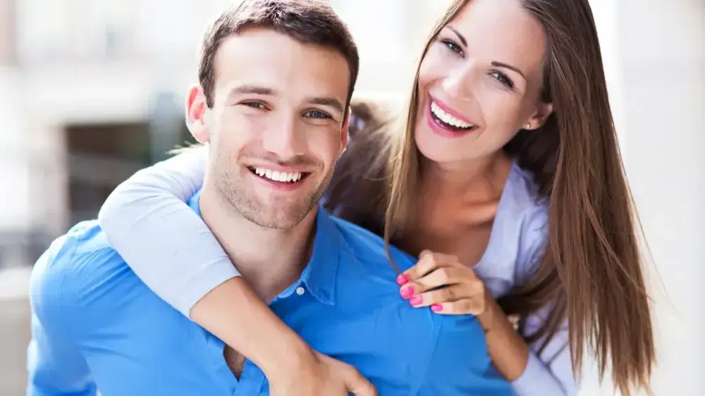 Ein Pärchen (Frau und Mann) lächelt in die Kamera, während die Frau den Mann umarmt.