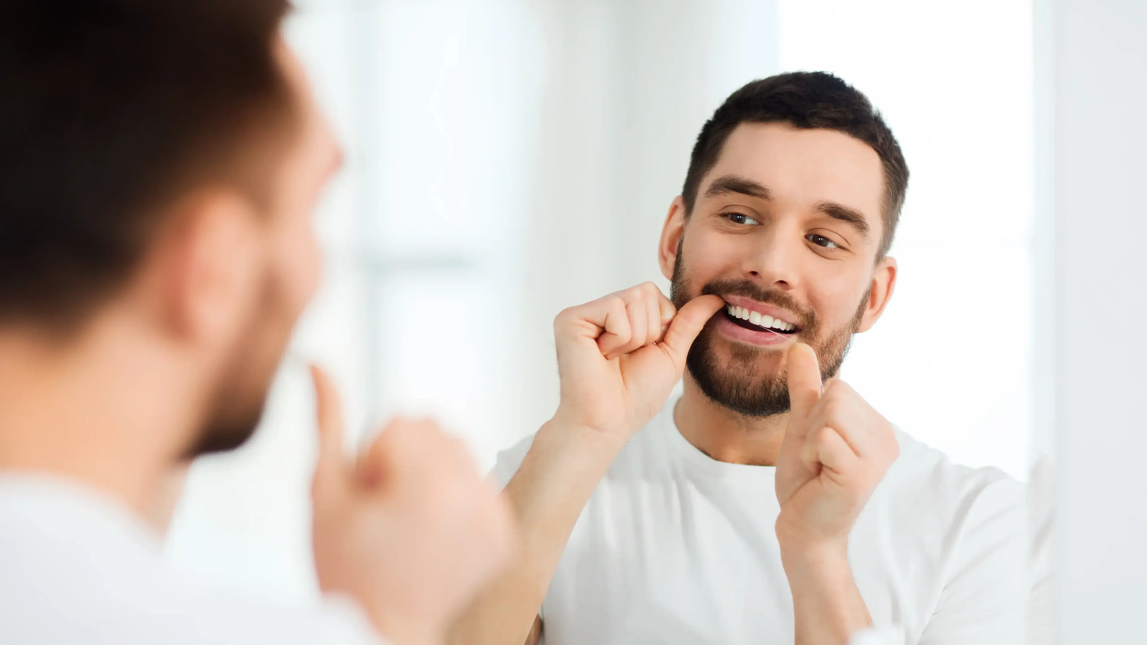 Ein junger Mann, der vor einem Badspiegel steht und Zahnseide nutzt.