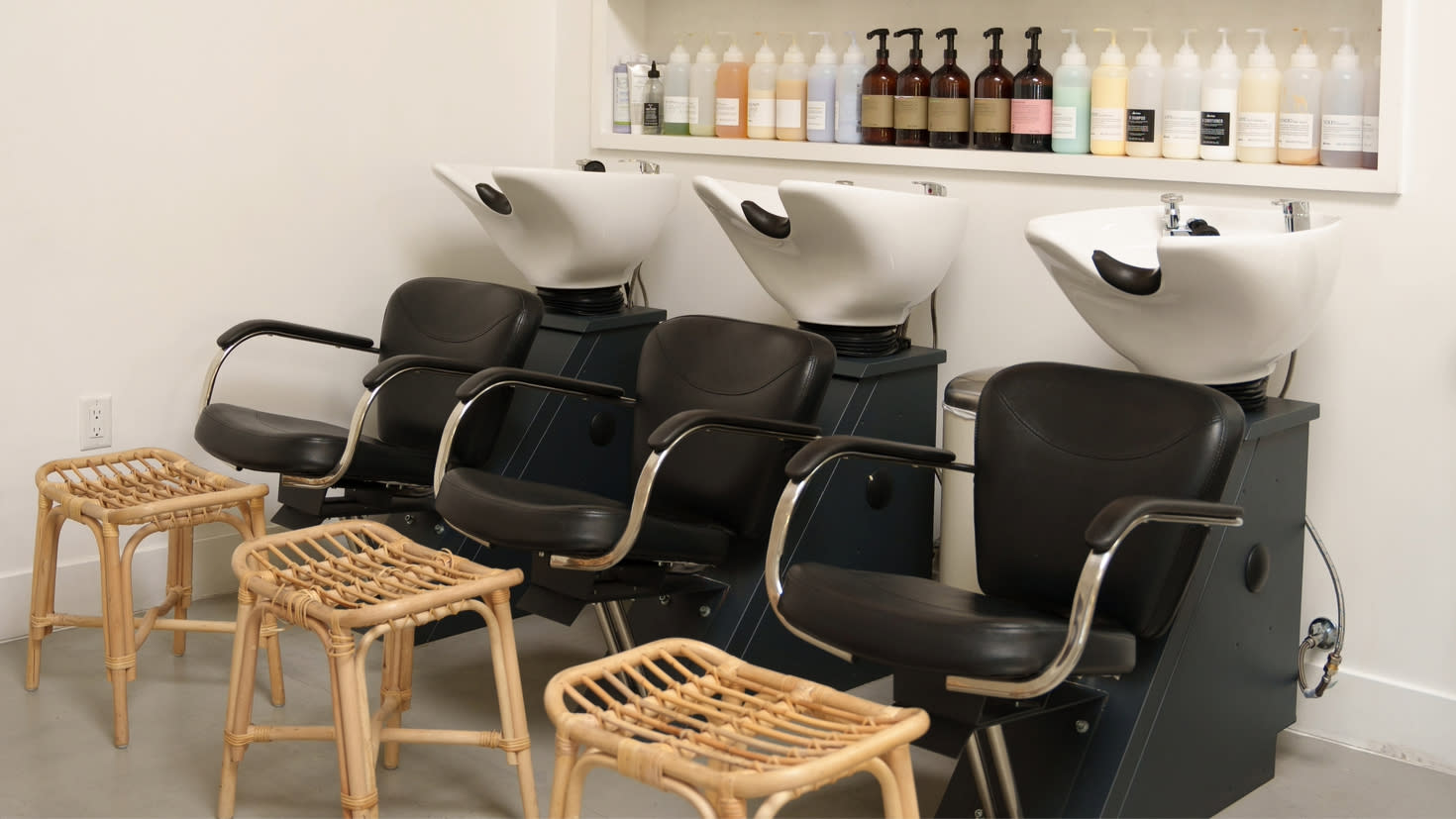 Salon hair washing station with 3 sinks, chairs, and stools in front of a wall with a shelf of hair products. 