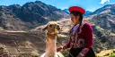 Woman in traditional clothing standing next to a llama, mountains in the backround.