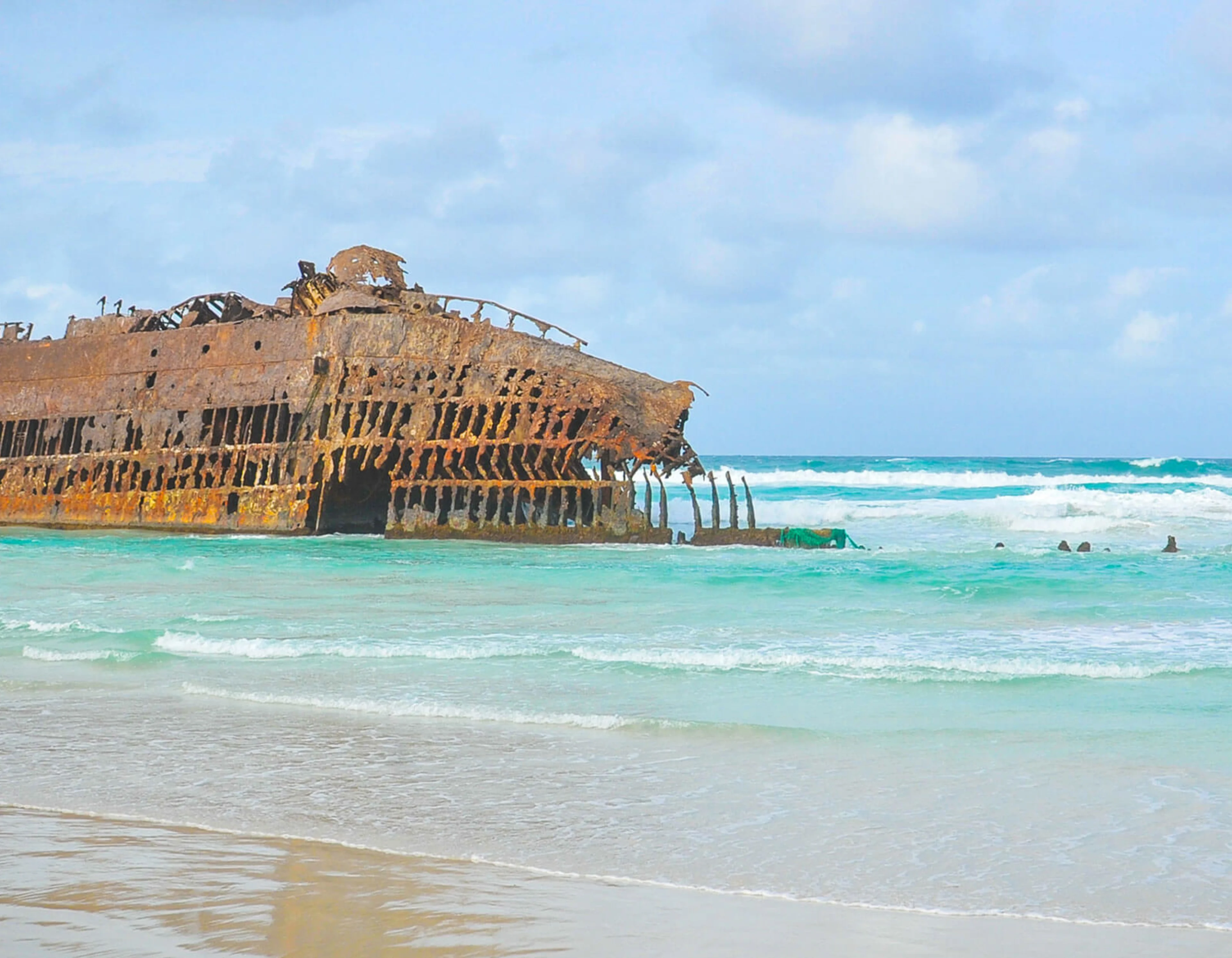 A large shipwreck is located at the water's edge on a beach. the sea is bright blue 