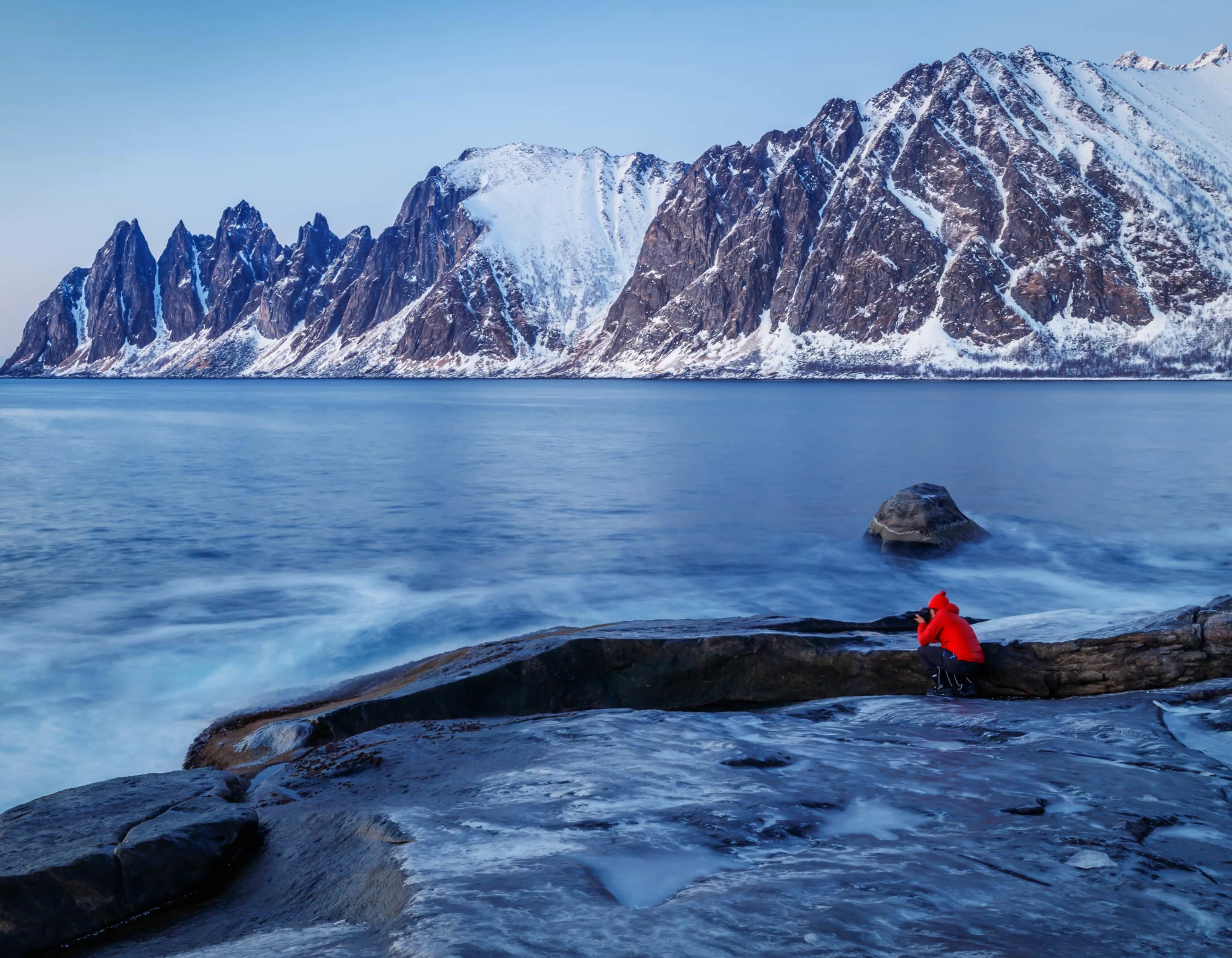 Senja, Norway, in winter. Person in a red jacket taking a picture of the sea, snow covered mountains in the background