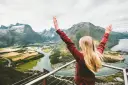 Person with blonde hair and arms up in the air looking out over fjord. 