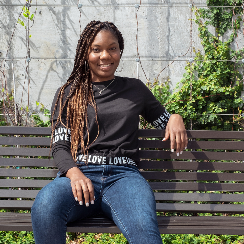 Black woman sitting on a bench and smiling, with plants in the background.