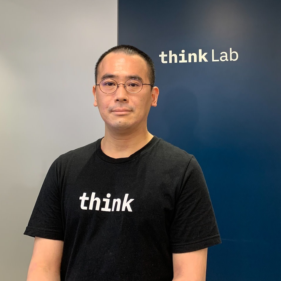 Man with glasses standing and smiling, with 'think' written on shirt and 'think lab' on wall