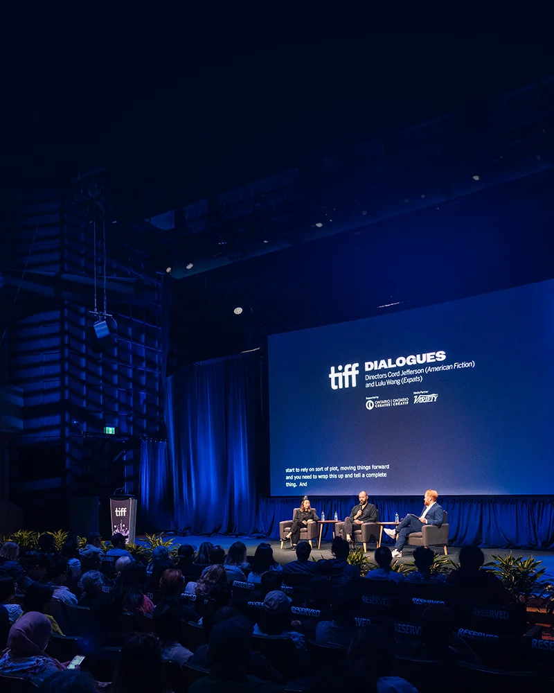 Three panelists on stage at a TIFF Dialogues event, speaking before a large audience in a blue-lit venue.