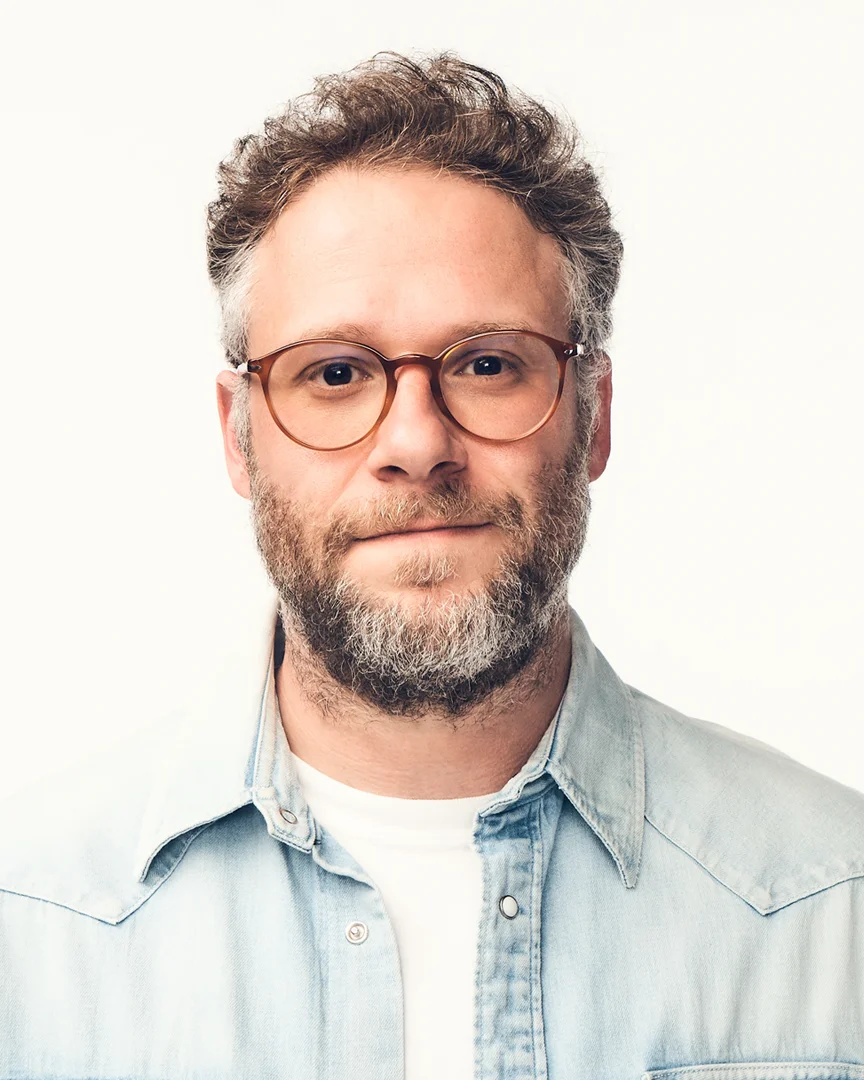Man with curly brown hair, round tortoiseshell glasses, and a beard wearing a light blue denim shirt.