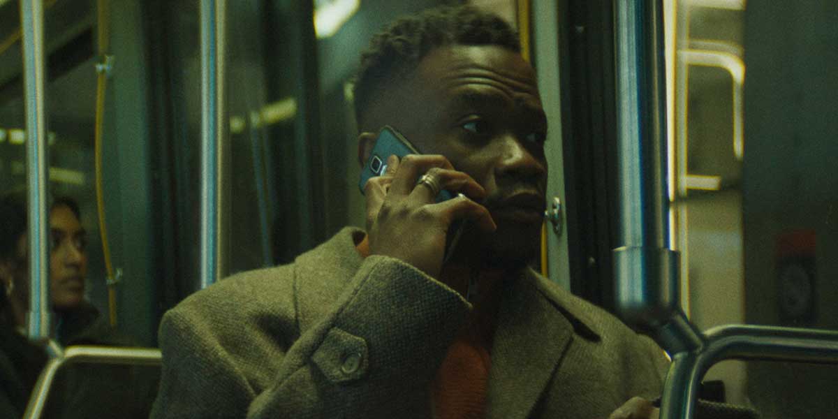 Person in olive coat talking on phone while riding public transit, dimly lit with greenish lighting