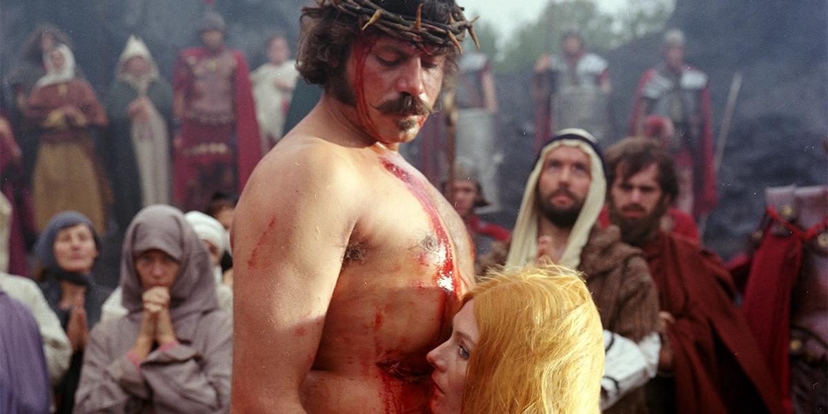 A shirtless, bloody man wearing a crown of thorns standing before a crowd as a woman leans closely against his chest.