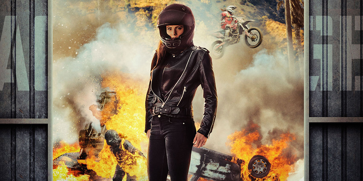 A stuntwoman dressed in black with a helmut standing in front of the Hollywood hills with explosions