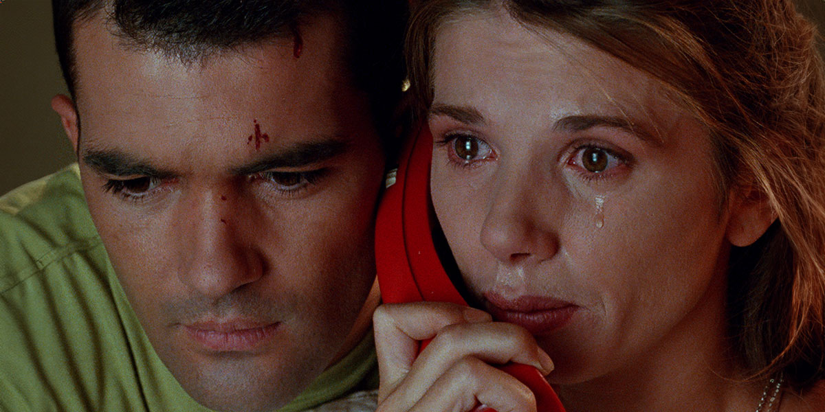 A woman listening to a red phone with a man beside her trying to listen as well from the otherside of the handset