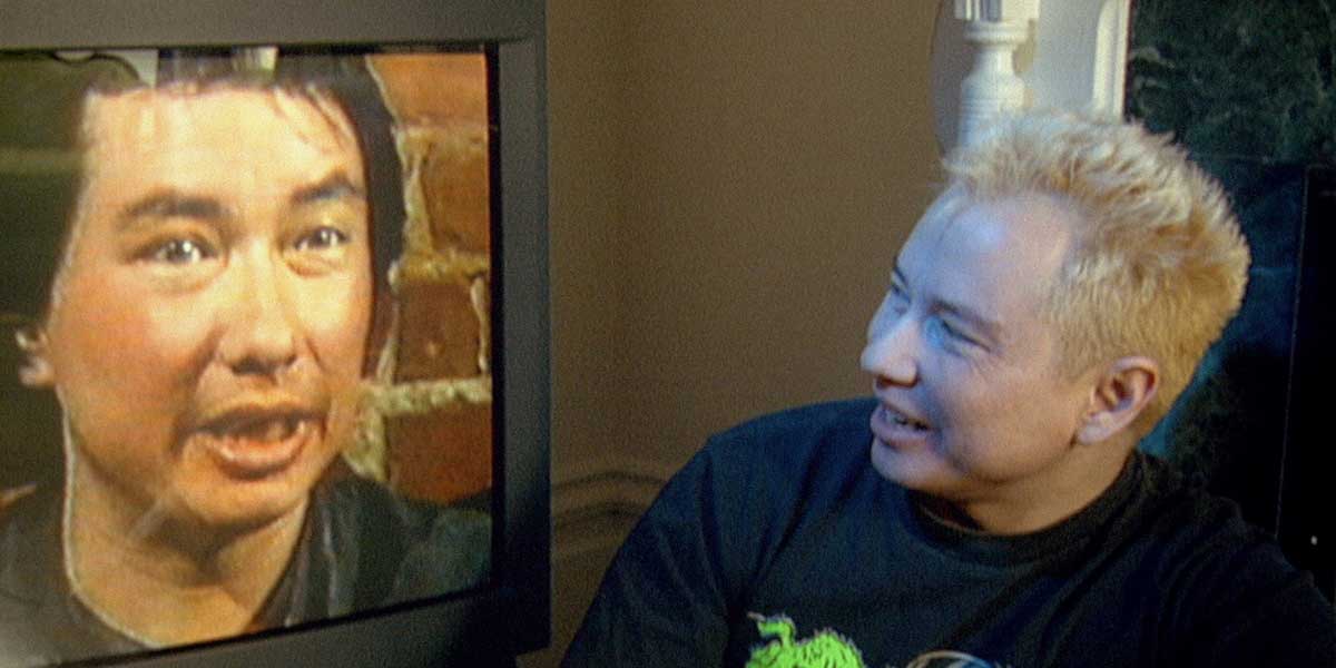 A person with blonde hair smiles while watching a face on a TV screen in a dimly lit room.
