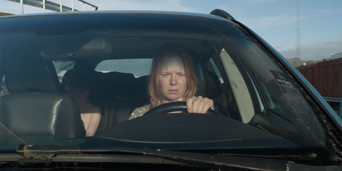 A women behind a steering wheel of a car squinting out onto the road with some kids behind her