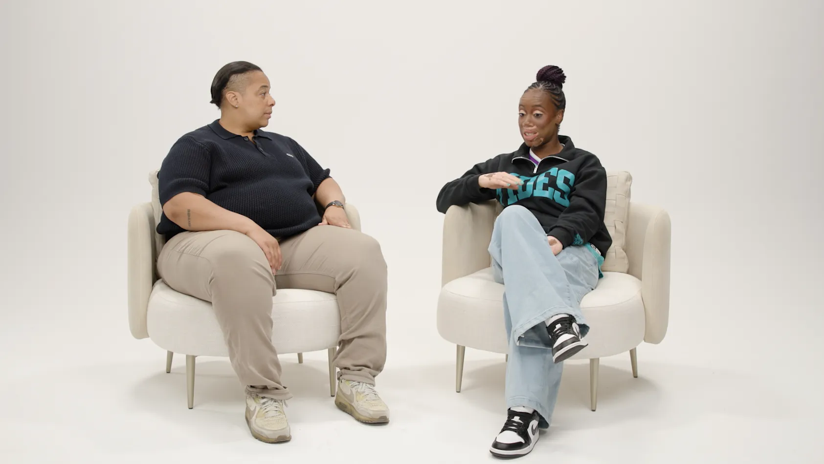 Two people sit in white chairs facing each other in conversation against a light background.