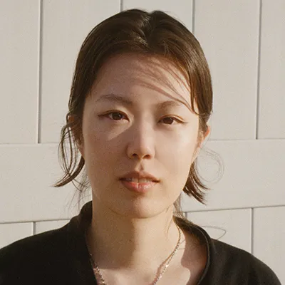 A woman with her hair tied back, wearing a black top standing against a white wall