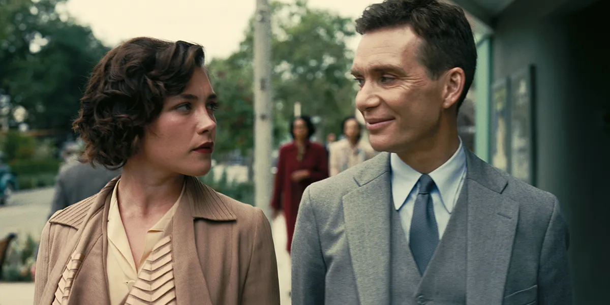 A man in a grey suit and a woman in a beige jacket share a conversation outdoors on a street.
