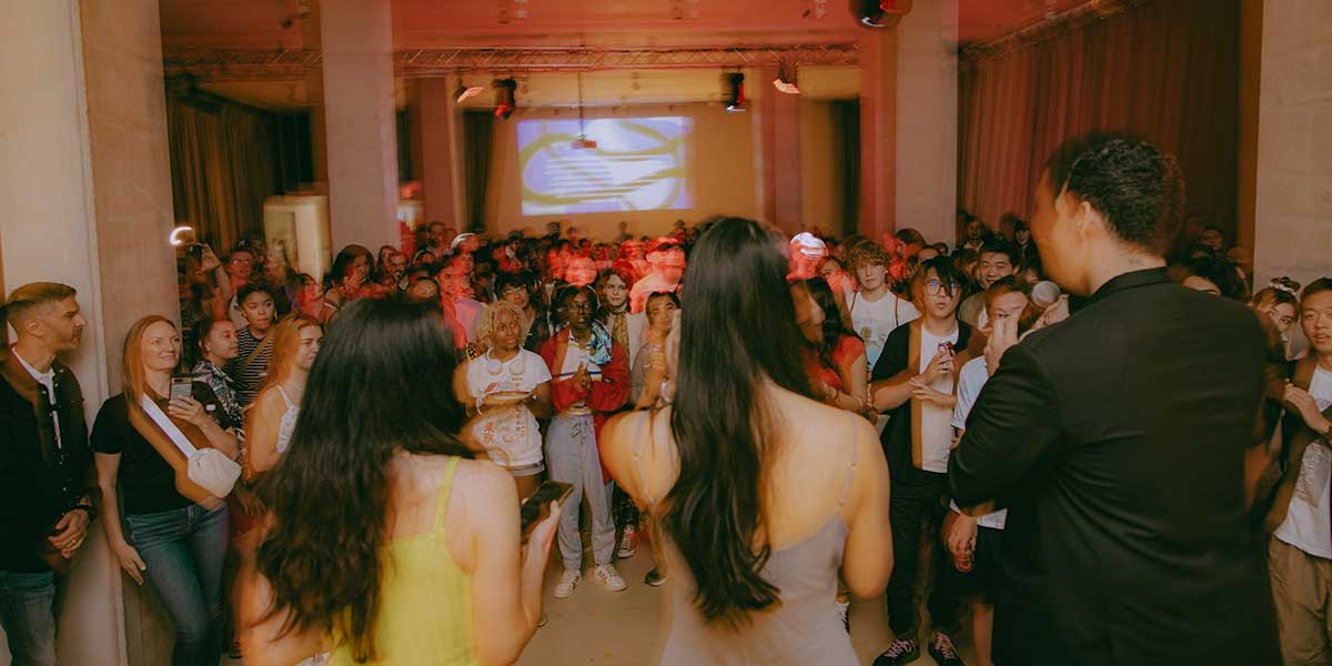Crowded indoor event space with red lighting, projection screen, and diverse audience watching performers on stage