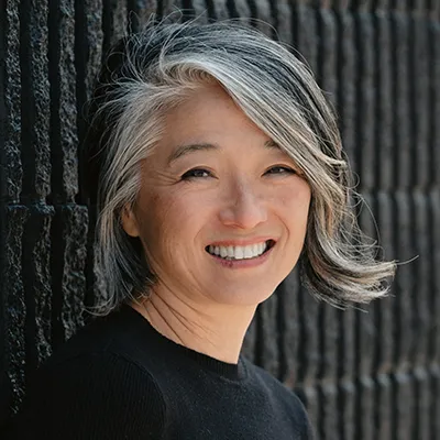 Smiling woman with silver-gray hair wearing black, photographed against dark striped background