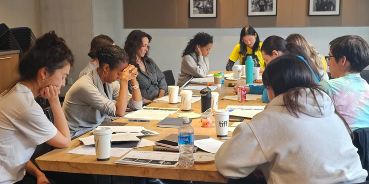 Students are seated at a long table studying documents during a workshop session, with coffee cups and papers visible