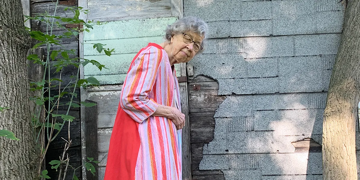 Elderly person in striped pink dress standing beside weathered wall and tree trunk in dappled sunlight