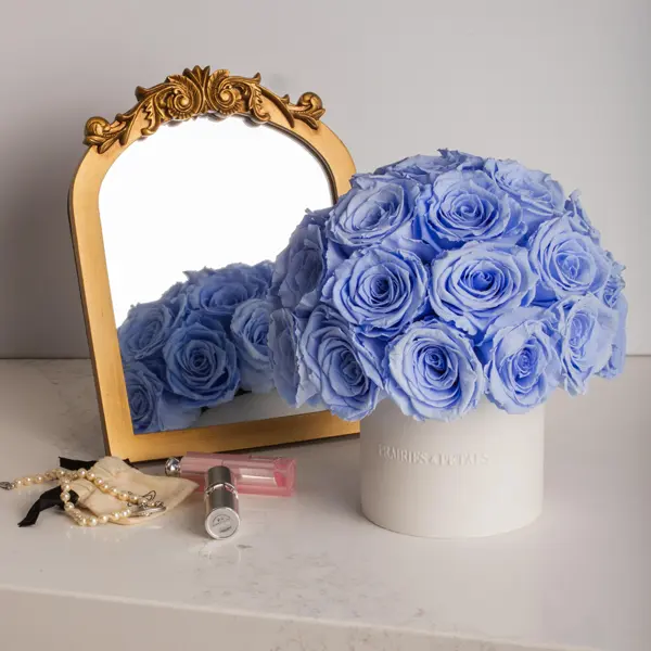 Light blue roses in white box next to ornate gold mirror with pearl necklace and perfume bottle on table