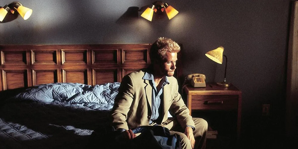 A man in a beige jacket sits pensively on a bed in a dimly lit motel room with warm lamp lighting.