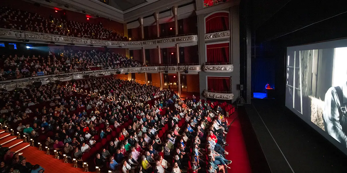 Packed theatre audience watching performance, with ornate balconies and red carpeting visible in dim lighting