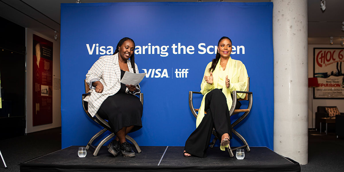 Sanaa Lathan sitting in a director's chair in front of the blue Visa backdrop talking to the audience