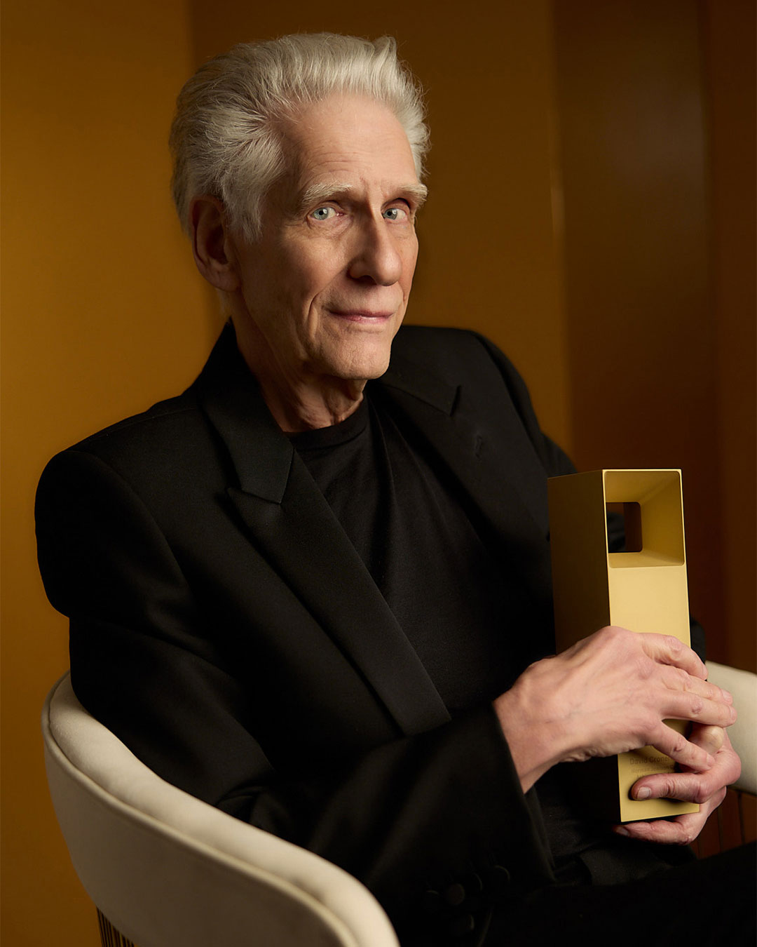 Portrait of David Cronenberg in a black suit sitting in a chair holding his award