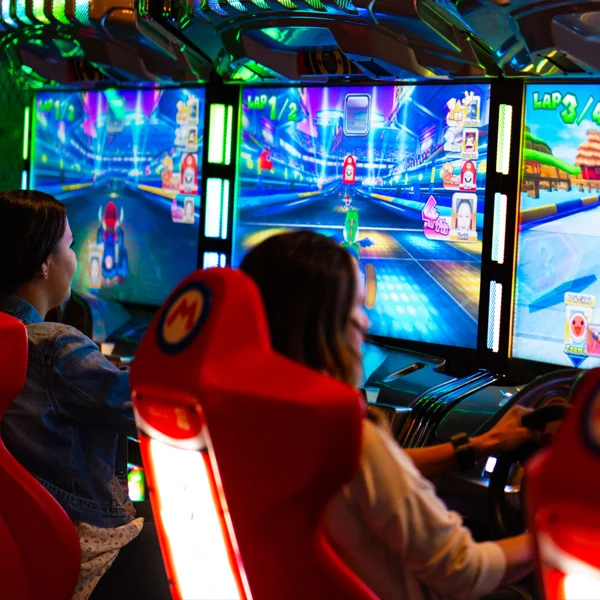 Two women playing the Mario Kart arcade game, seated in red racing seats with glowing screens.