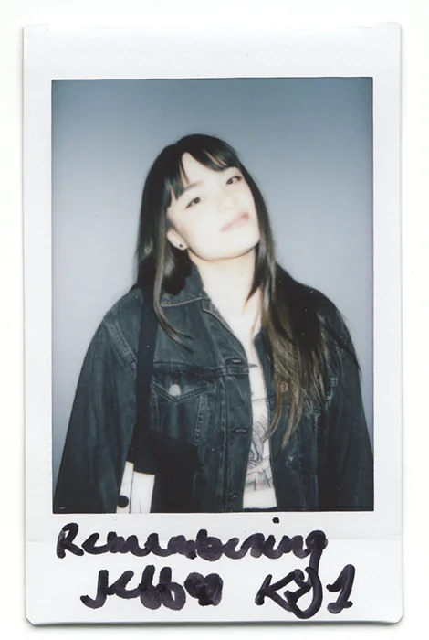 Polaroid photo of a young woman with long dark hair in a denim jacket, text reads "Remembering Kebo KS1"