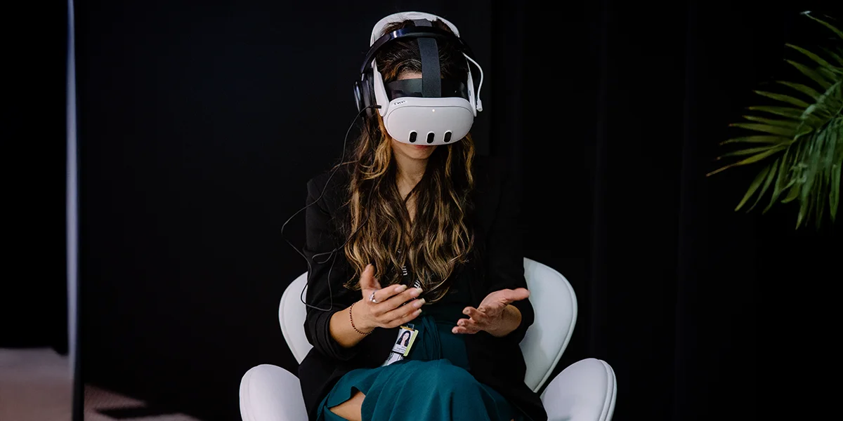 Woman sitting in a white chair wearing a VR headset, gesturing with hands, dark background.
