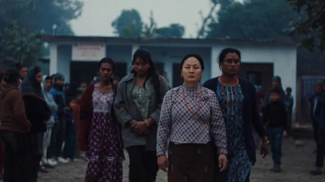 A group of women walk forward in a misty village setting, with a white building and crowd behind them.