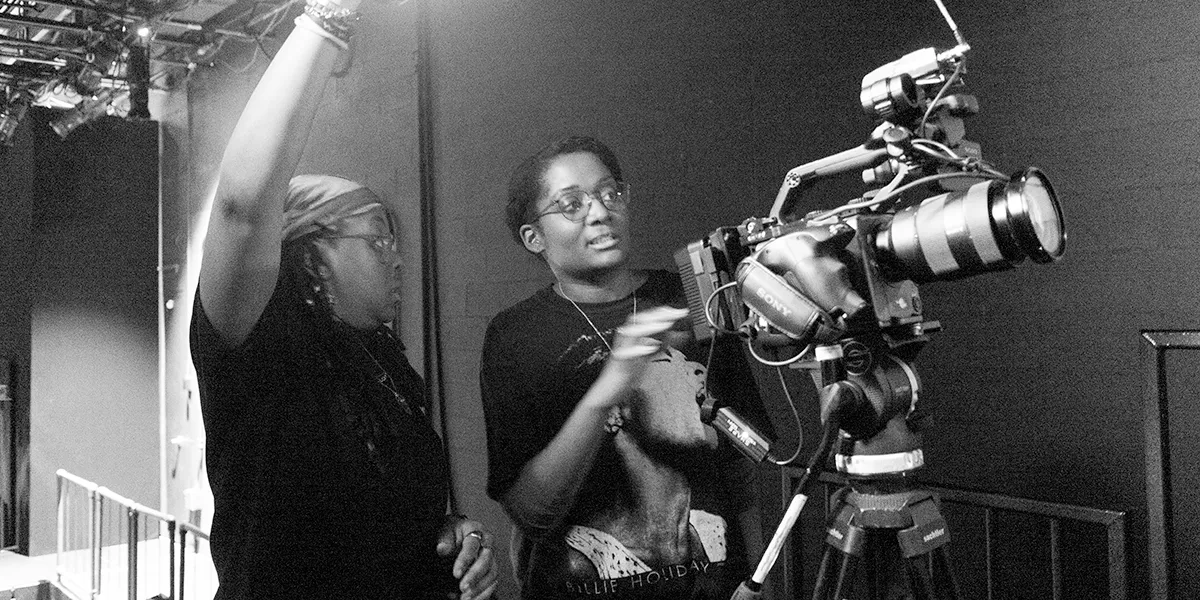 Two people working on a film set, one pointing at a light while the other gestures near a camera.