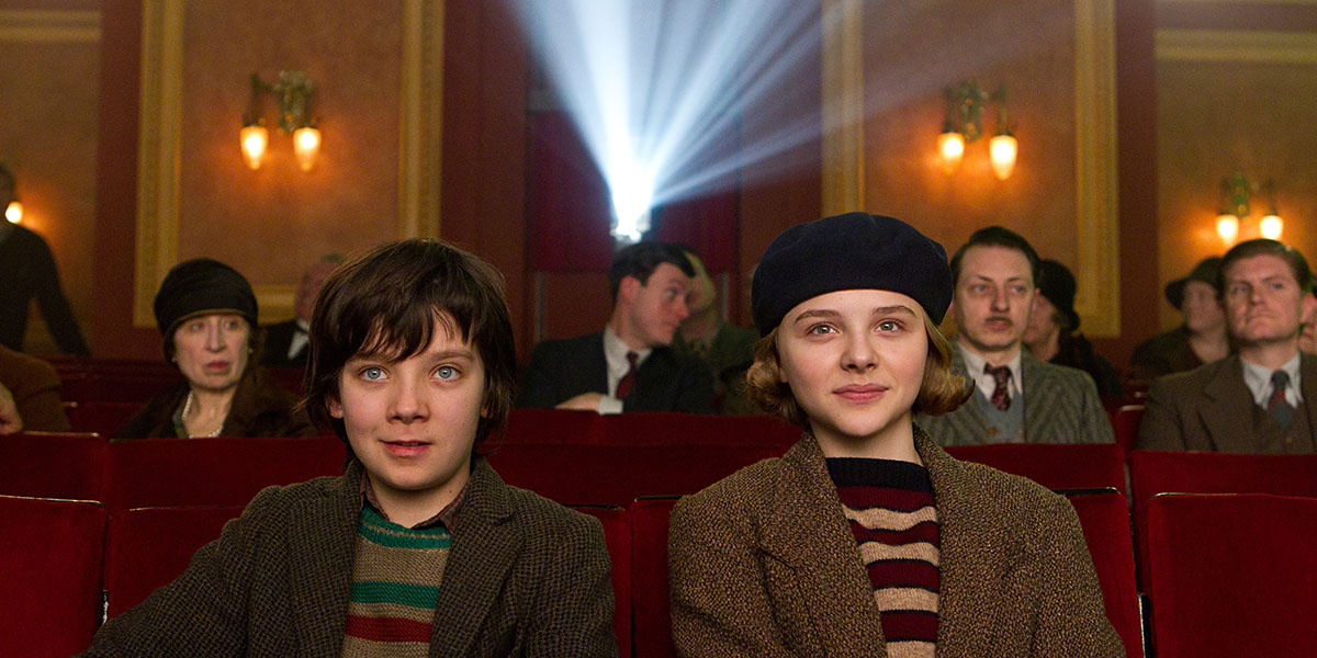 Hugo (Asa Butterfield) and Isabelle (Chloë Grace Moretz) sitting in a theatre watching the screen as a projector runs behind them