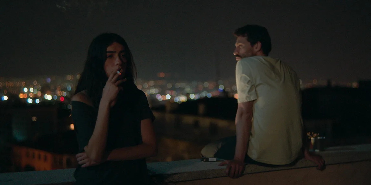 Two people smoking at night on a rooftop with city lights twinkling in the dark background