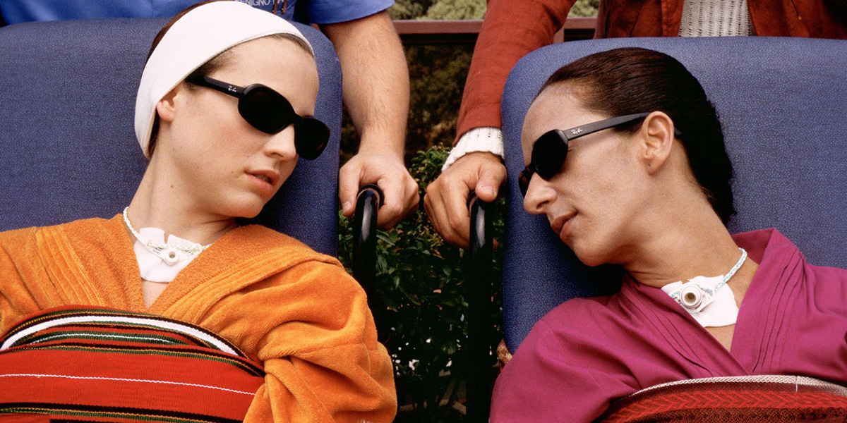 Two woman in bathrobes and sunglasses looking at each other as they sit in wheelchairs and being pushed by other people