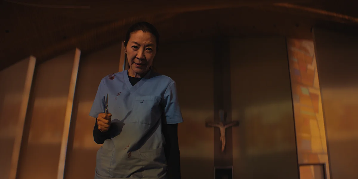 A woman in blood-stained blue scrubs holds scissors in a dimly lit room with a crucifix behind her.