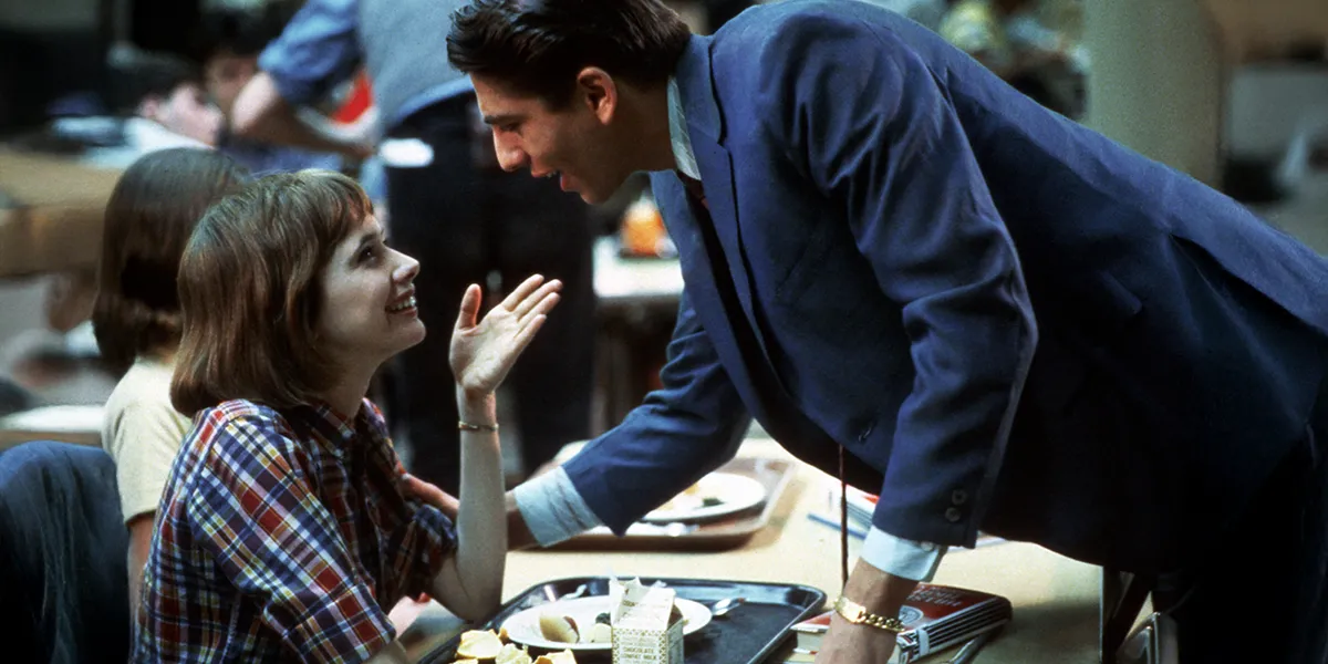 A young woman in plaid smiles up at a man in a blue suit leaning over a cafeteria table.
