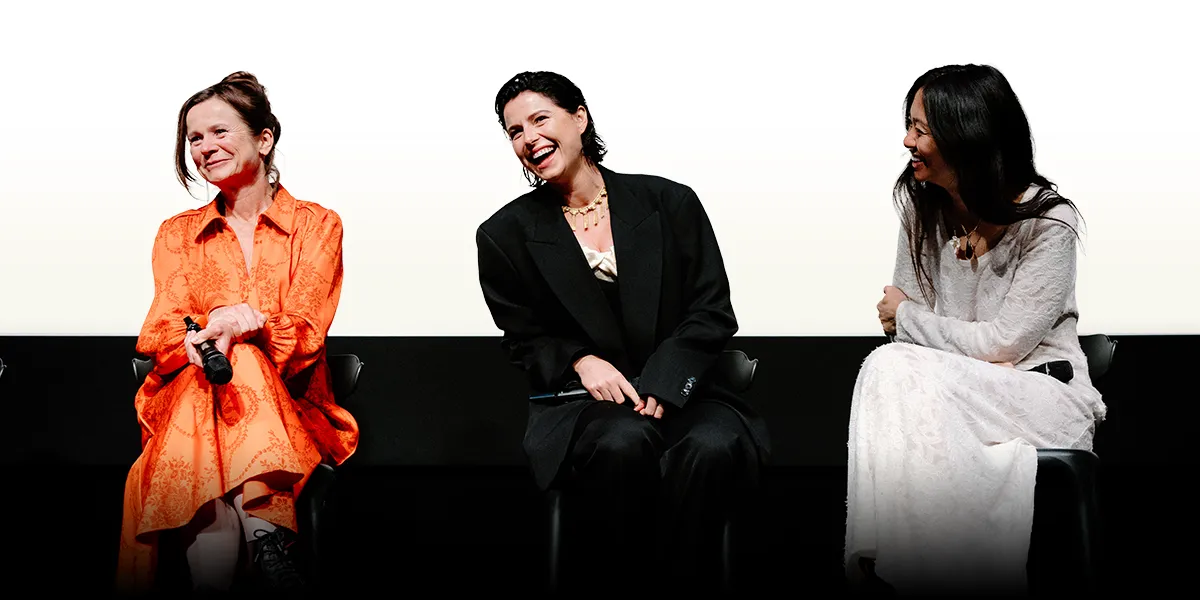 Three women sit laughing on stage during a panel, dressed in orange, black, and white outfits.