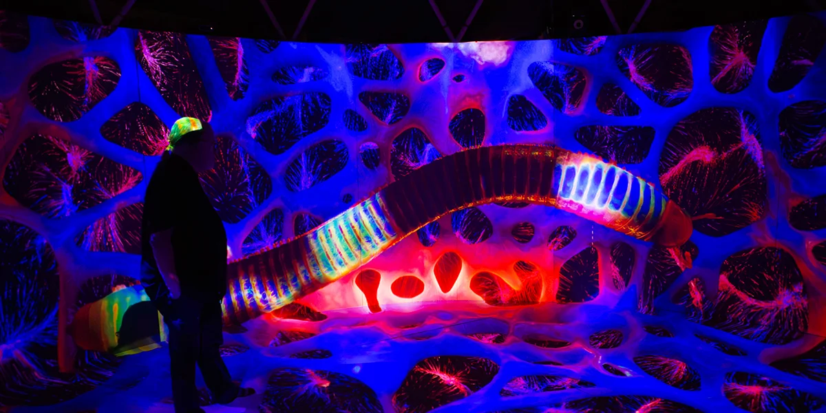 A person stands in a vivid UV-lit immersive art installation with glowing blue, red, and neon organic shapes.