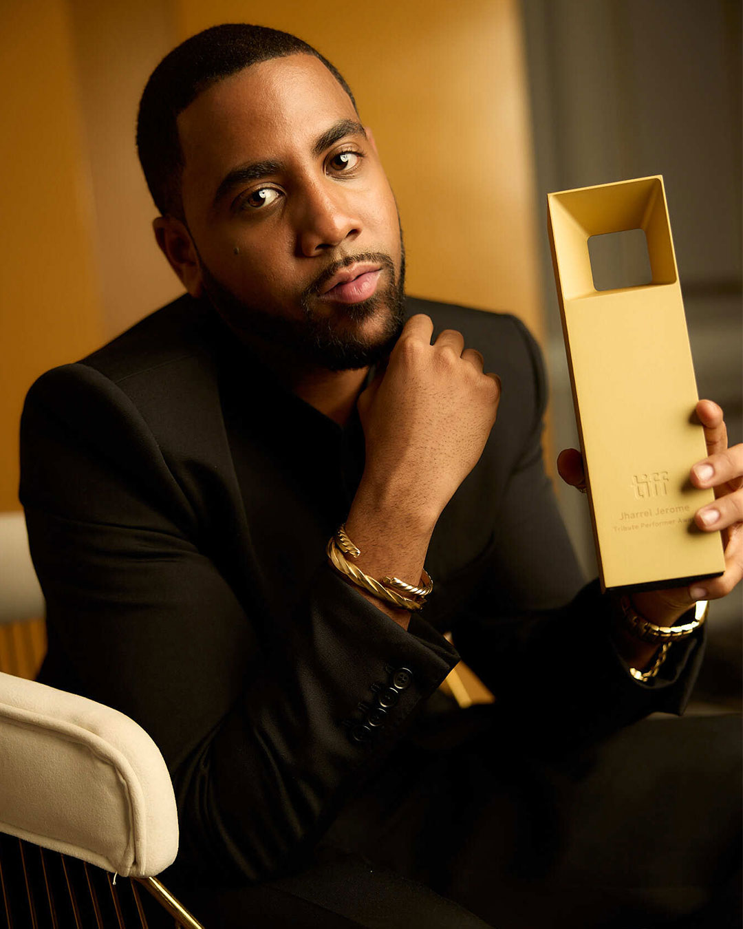 Portrait of Jharrel Jerome in a black suit sitting in a chair and holding his award