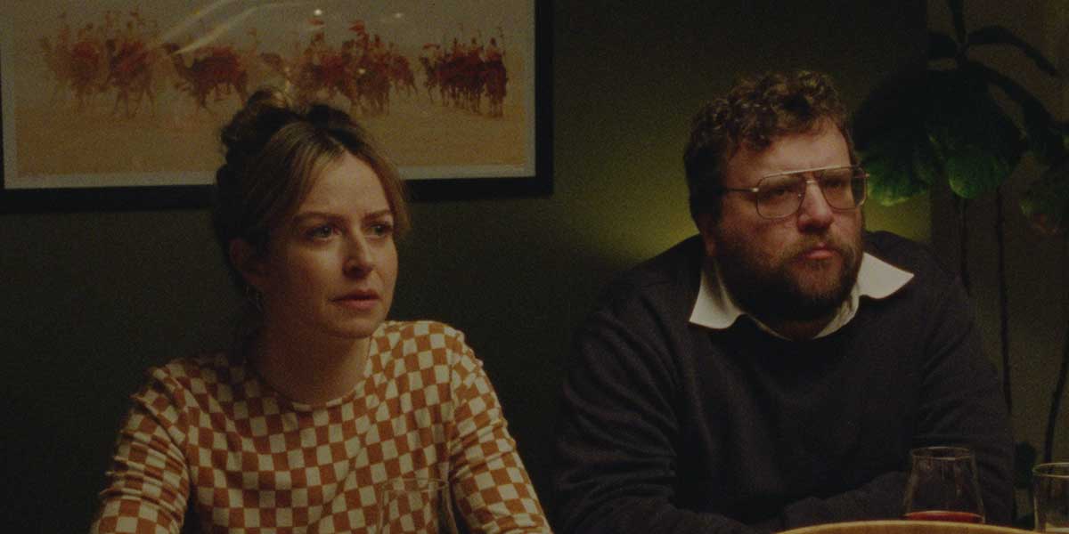 Two people sitting at a dimly lit table, woman in checkered sweater and man in dark clothing with glasses