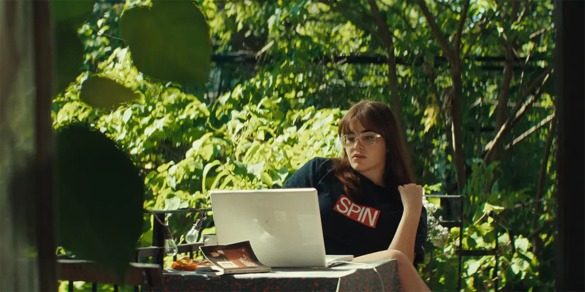 A person wearing glasses and a SPIN t-shirt works on a laptop outdoors surrounded by green foliage