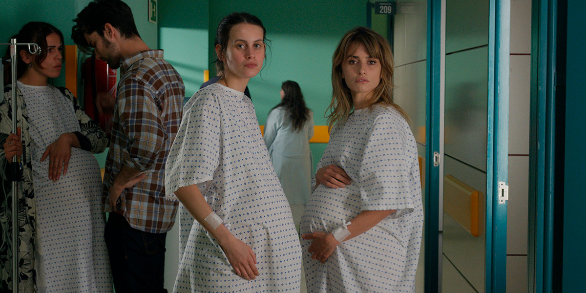 Two pregnant women in hospital gowns facing one another but both looking towards the camera