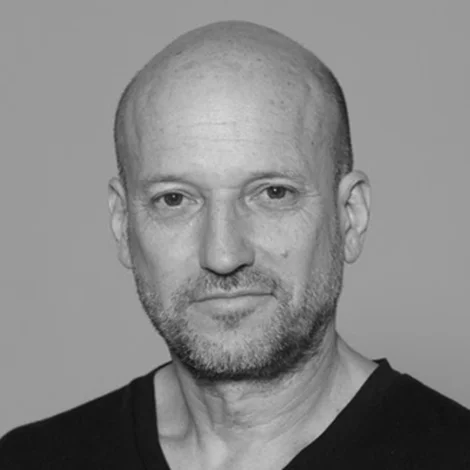 Black and white headshot of a bald middle-aged man with stubble, wearing a dark v-neck shirt.